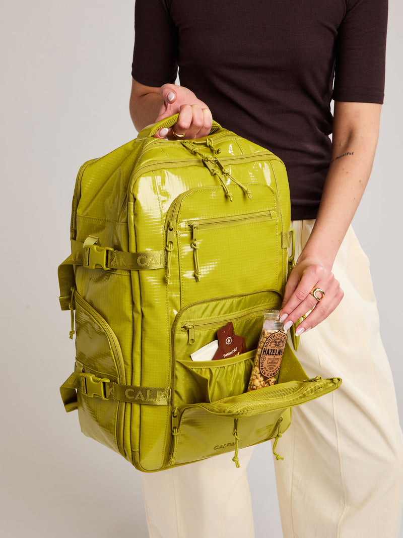 Person adding snacks to the front pocket of the CALPAK Terra 26L Laptop Backpack in yellow green Wasabi with multiple pockets on a white background