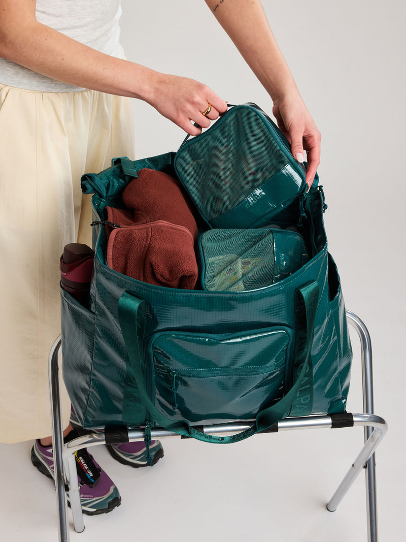 A dark teal green Kale travel  CALPAK Terra 35L Tote bag with a front zippered pocket, being packed with toiletry bags by a person