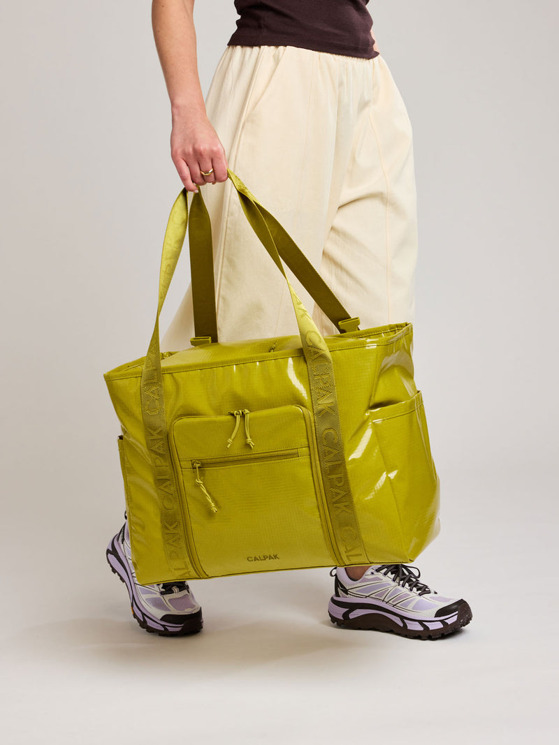 A person wearing cream-colored pants and athletic shoes holds a large, shiny lime green Wasabi CALPAK Terra 35L Laptop tote bag with dual shoulder straps against a plain light gray studio background