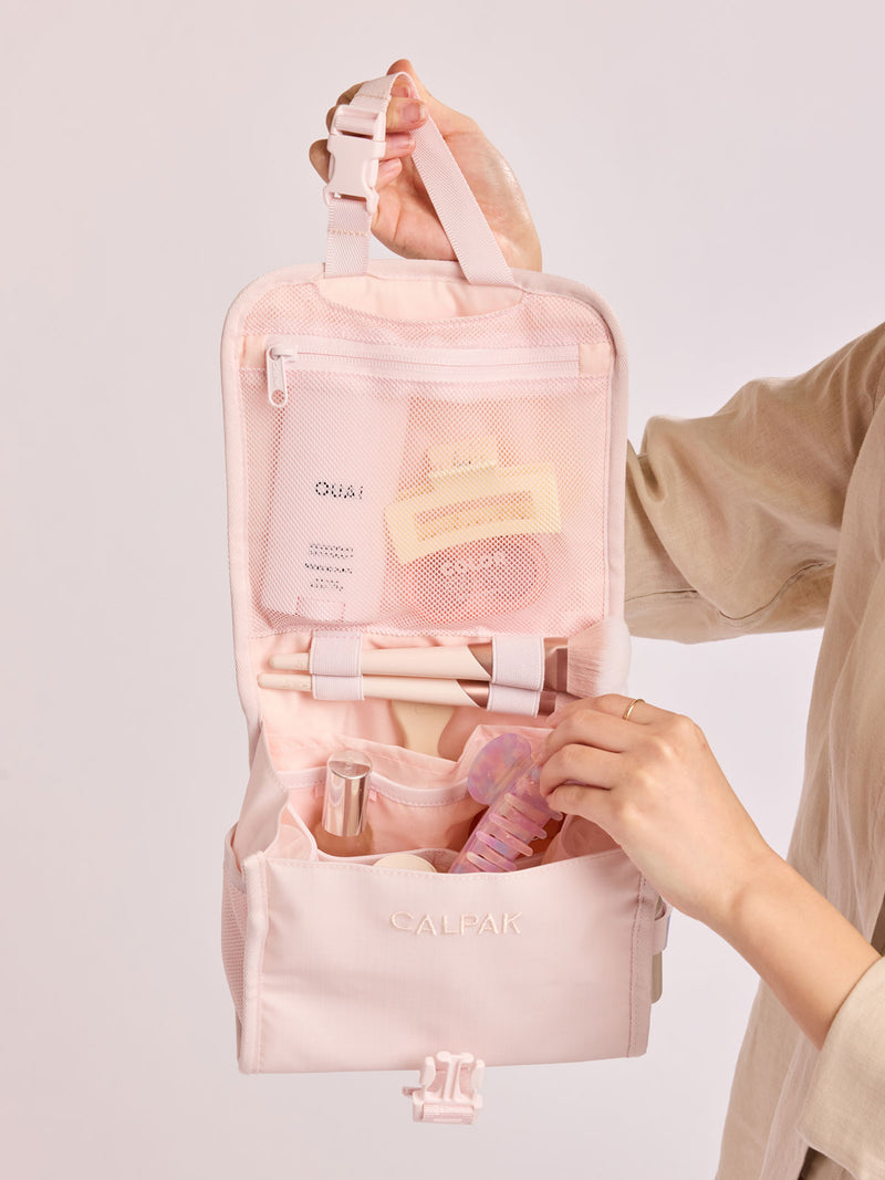 Putting toiletry items into CALPAK Terra Hanging Toiletry Bag in light pink Shell Pink with front buckle