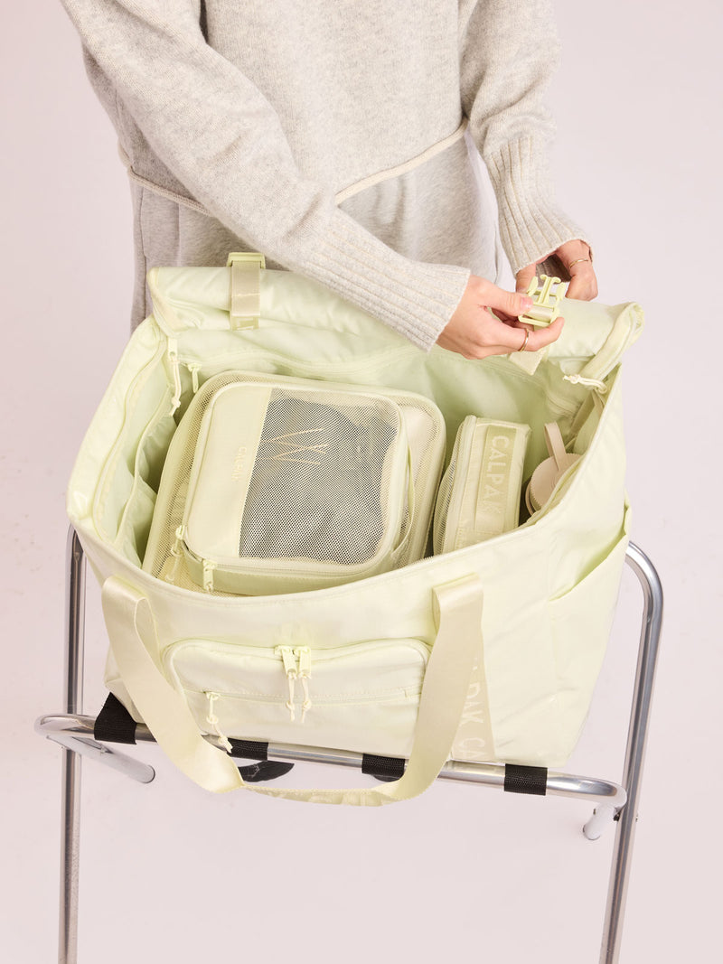 Model adding the small and medium packing cubes of CALPAK Terra 5-Piece Packing Cube Set in light green Celadon with two small cubes, two medium cubes, and one large mesh cube with zippers and mesh into a bag