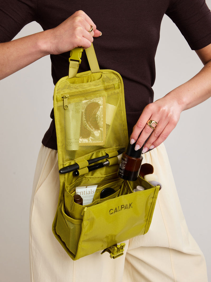 Model adding toiletry into CALPAK Terra Hanging Toiletry Bag to store makeup and toiletries in yellow green Wasabi on a white background