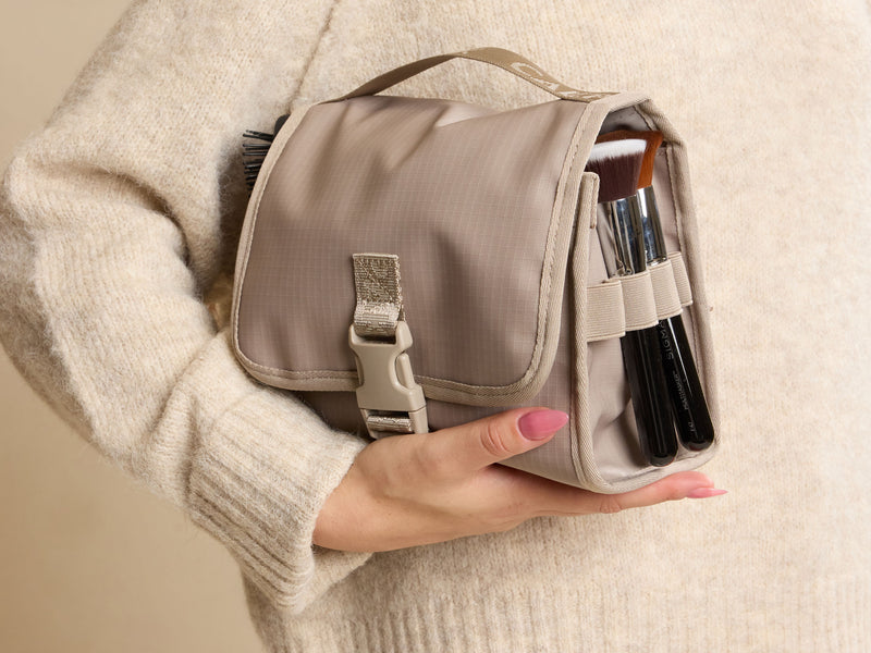 Person holding a beige Terra Hanging Toiletry Bag with makeup brushes inside against a beige background