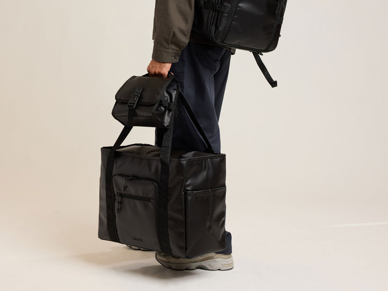 Person holding a CALPAK Terra black bag with multiple compartments on a beige background