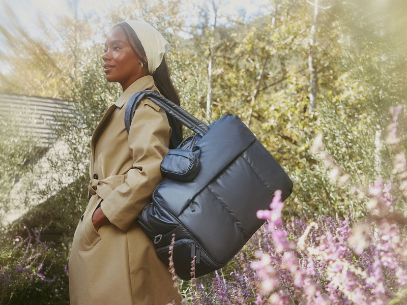 Woman holding a metallic indigo CALPAK travel bag outdoors with greenery and flowers in the background