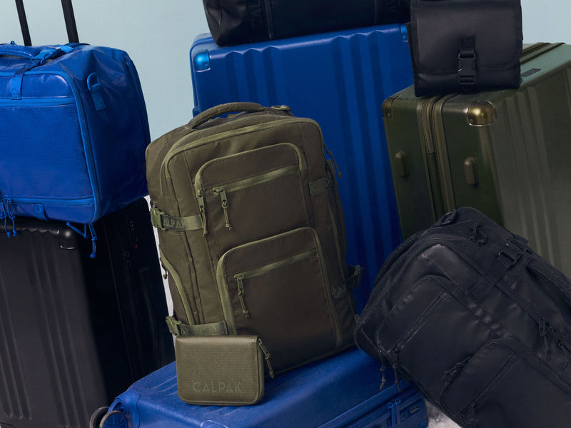 Green, blue and black backpacks with visible brand logo on a pile of suitcases and bags from CALPAK.