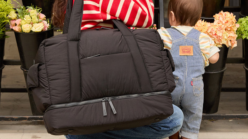 Person holding a black CALPAK Weekender Duffel bag with a child in overalls sitting next to them, surrounded by potted plants.