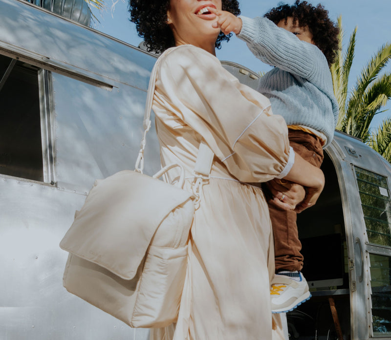 Woman in a beige outfit holding a child in front of a vintage trailer with palm trees in the background while wearing a CALPAK Convertible Mini Diaper Bag  in beige Oatmeal