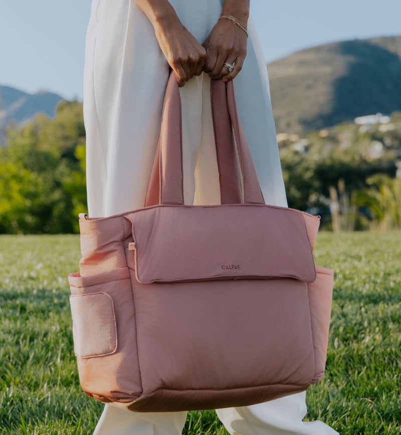 Person holding a CALPAK Diaper Tote Bag in pink Peony outdoors with greenery and mountains in the background