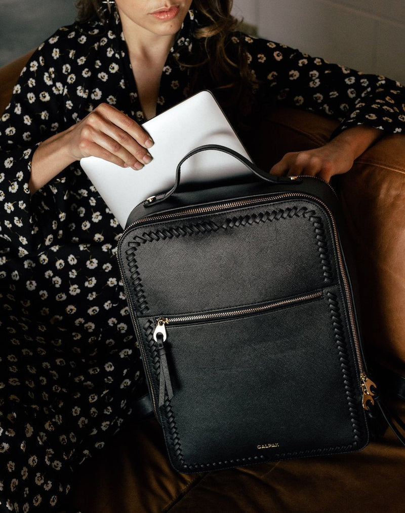 The Kaya Laptop Backpack in black, shown by a model holding the bag as she slips a laptop into the rear compartment